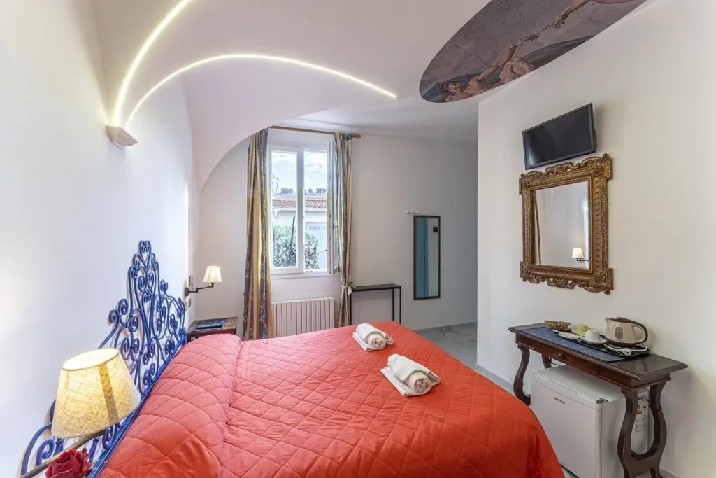 Camera d'hotel a tema progettata dallo studio di progettazione hotel AlitiniDesign, ispirata a Michelangelo. La stanza presenta una testata letto in ferro battuto blu, un soffitto a volta con dettagli illuminati a LED e un affresco della Creazione di Adamo. Arredi eleganti, con specchio dorato e un tavolino da tè completano l'ambiente accogliente e raffinato.