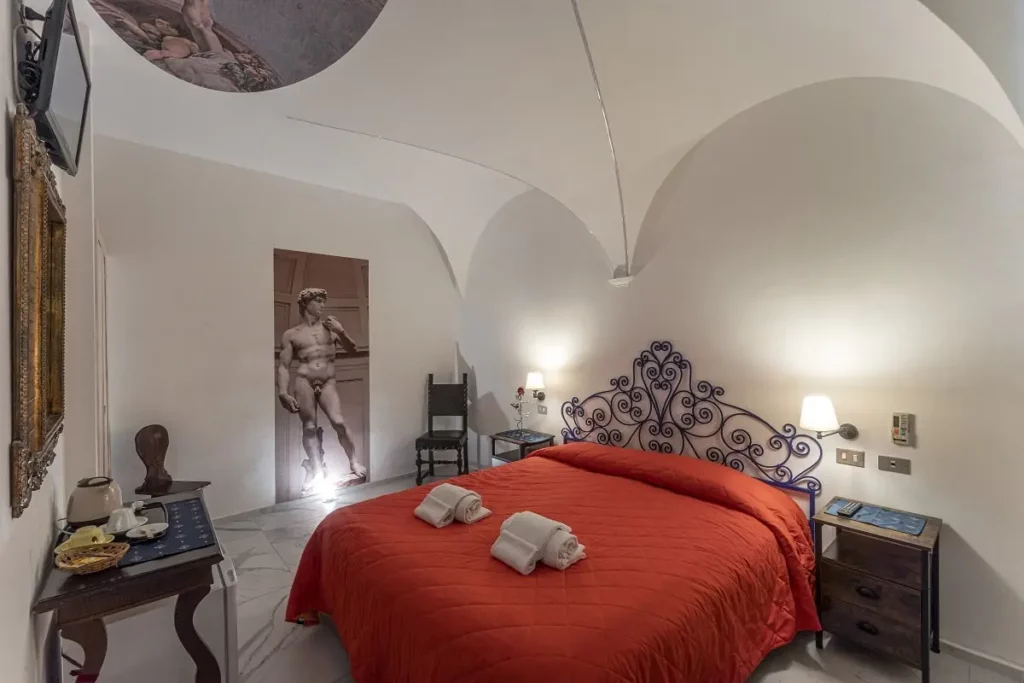 Camera d'hotel a tema progettata dallo studio di progettazione hotel AlitiniDesign, ispirata a Michelangelo. La stanza presenta una testata letto in ferro battuto blu, un soffitto a volta con dettagli illuminati a LED e un affresco della Creazione di Adamo. Arredi eleganti, con specchio dorato e un tavolino da tè completano l'ambiente accogliente e raffinato.