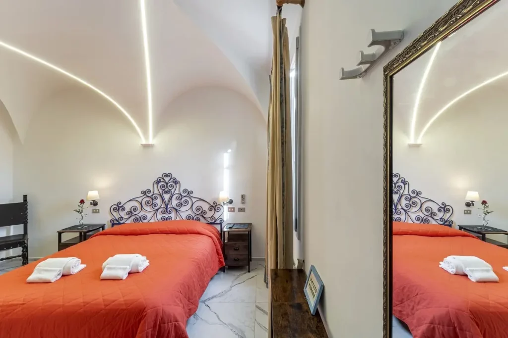 Camera d'hotel a tema progettata dallo studio di progettazione hotel AlitiniDesign, ispirata a Michelangelo. La stanza presenta una testata letto in ferro battuto blu, un soffitto a volta con dettagli illuminati a LED e un affresco della Creazione di Adamo. Arredi eleganti, con specchio dorato e un tavolino da tè completano l'ambiente accogliente e raffinato.