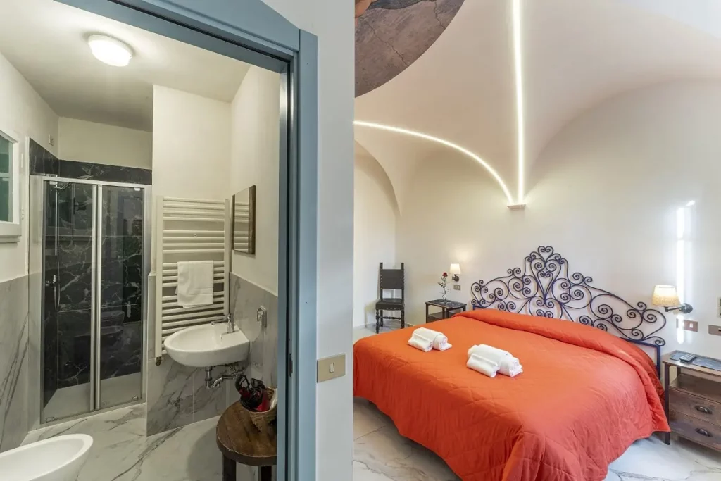 Camera d'hotel a tema progettata dallo studio di progettazione hotel AlitiniDesign, ispirata a Michelangelo. La stanza presenta una testata letto in ferro battuto blu, un soffitto a volta con dettagli illuminati a LED e un affresco della Creazione di Adamo. Arredi eleganti, con specchio dorato e un tavolino da tè completano l'ambiente accogliente e raffinato.